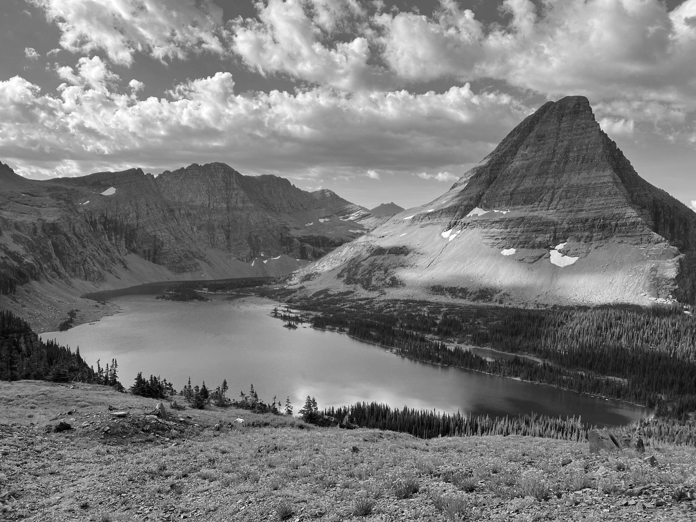 A still alpine lake set beneath weathered mountain peaks under a wide, clouded sky.